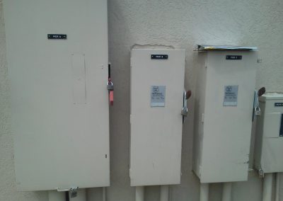 Three beige electrical panels with labels on a textured wall; two panels have padlocks on their handles.