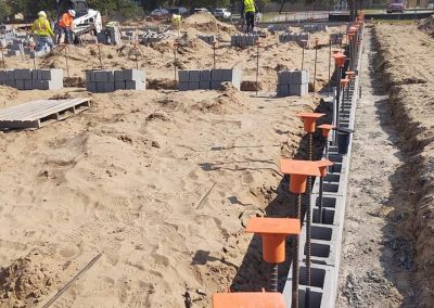 Construction site with concrete blocks, metal rods, sand, and workers in safety vests under a partly cloudy sky.
