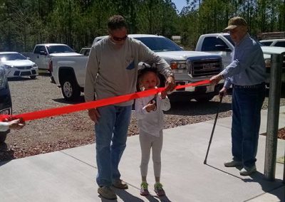 A young girl cuts a red ribbon at an outdoor event, with two men assisting her. Cars are parked in the background.