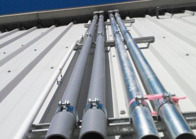 View looking up at several metal and PVC pipes attached to the side of a corrugated building against a blue sky.