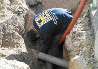 A worker in a trench installs or repairs pipes in a construction area, wearing a blue shirt with yellow text.