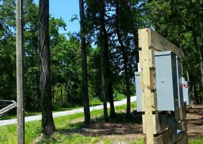 Wooden panel with electrical boxes near trees, next to a pole with a streetlight on a sunny day.
